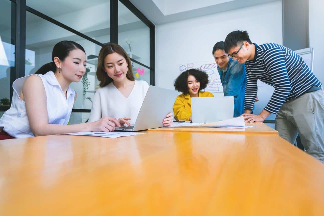 A group of young Asian men and women sitting at a conference desk and referring to their laptops and having a discussion, representing first-time entrepreneurs brainstorming.